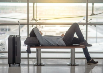 Young Asian man with suitcase luggage and backpack lying on bench in airport terminal