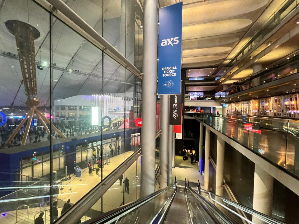 A view looking down the escalator at THe O2. the priority entrance is visible at the bottom and a crowd of people beyond.