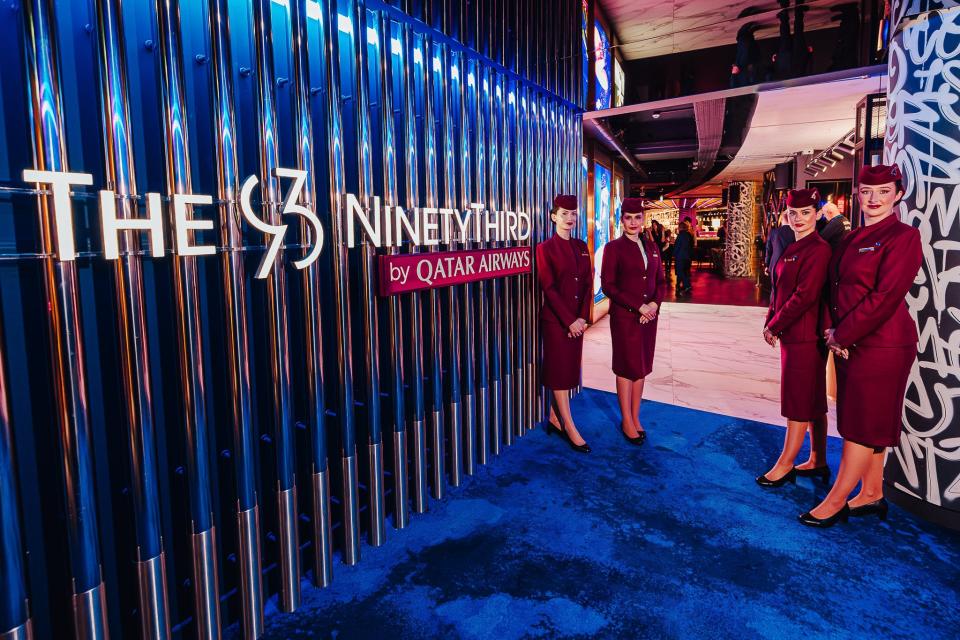 Four Qatar Airways flight attendants wearing red uniforms stand outside the entrance sign to The NinetyThird