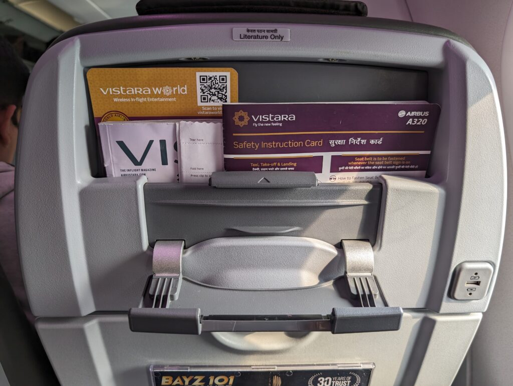 The back of a Vistara economy class seat with safety information cards and a built in tablet holder.