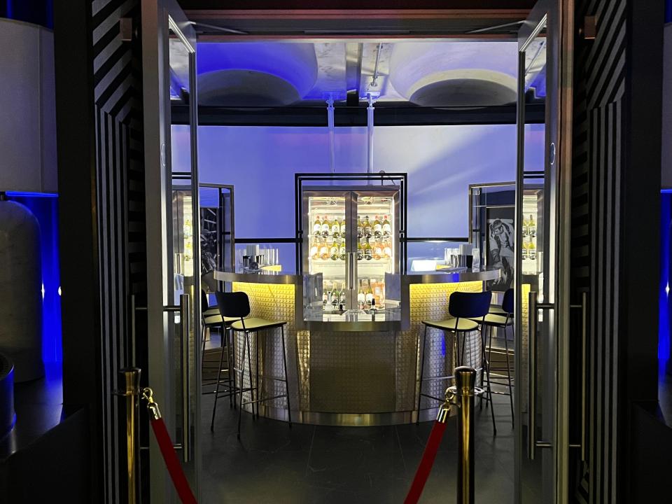 A bar on a horseshoe-shaped balcony has four stools and a large wine fridge in the rear. A velvet rope blocks the entrance during the concert.