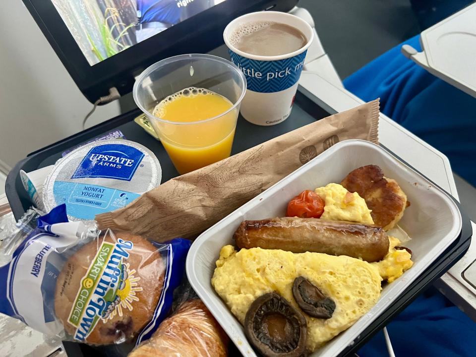 The breakfast: eggs sausage, muffin, yogurt, juice, coffee, and a croissant on a tray.