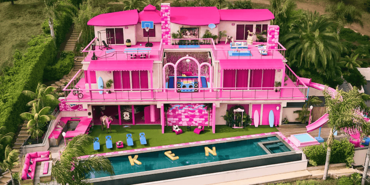 A vibrant, Barbie-themed pink mansion with a pool spelling "KEN" is surrounded by lush greenery, featuring various outdoor play areas and elaborate decorations.