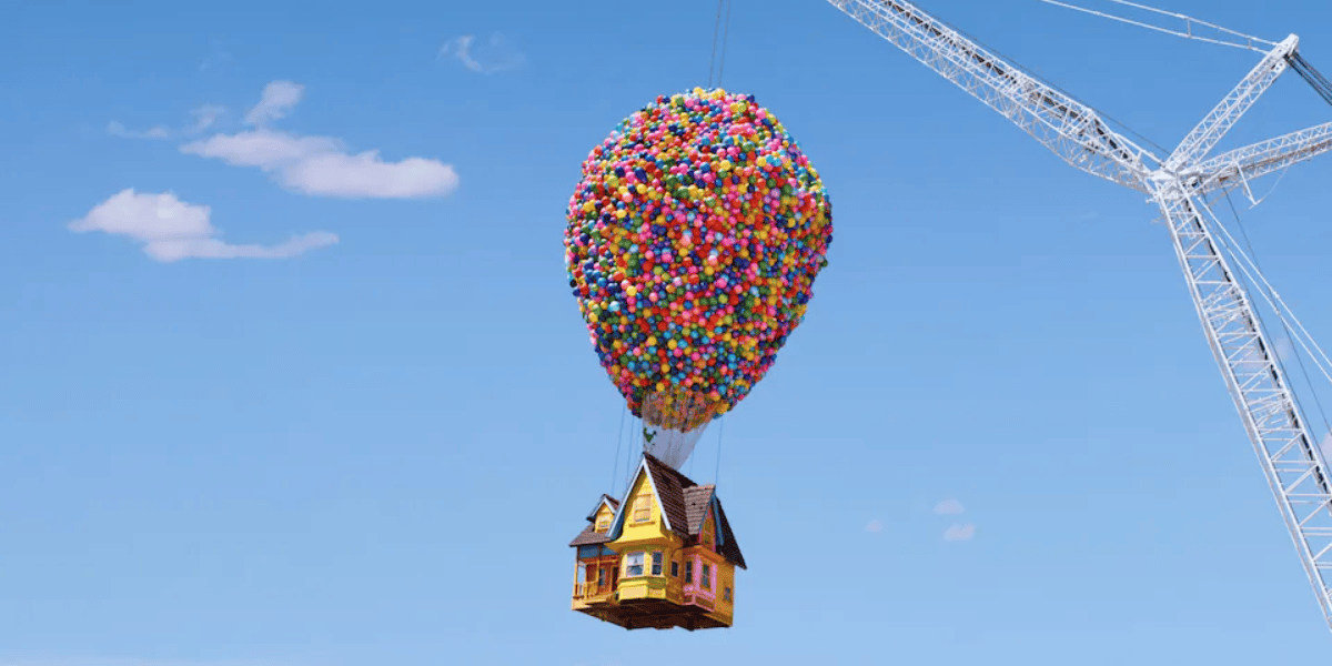 A colorful floating house, lifted by balloons, drifts in a clear blue sky beside a construction crane, reminiscent of a scene from the animated movie "Up"—now an UP-Inspired Airbnb.