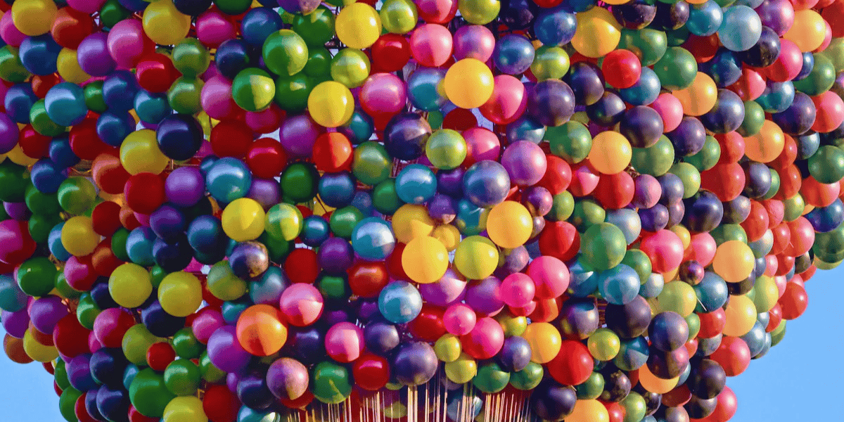 A massive cluster of colorful balloons fills the frame against a clear blue sky background.
