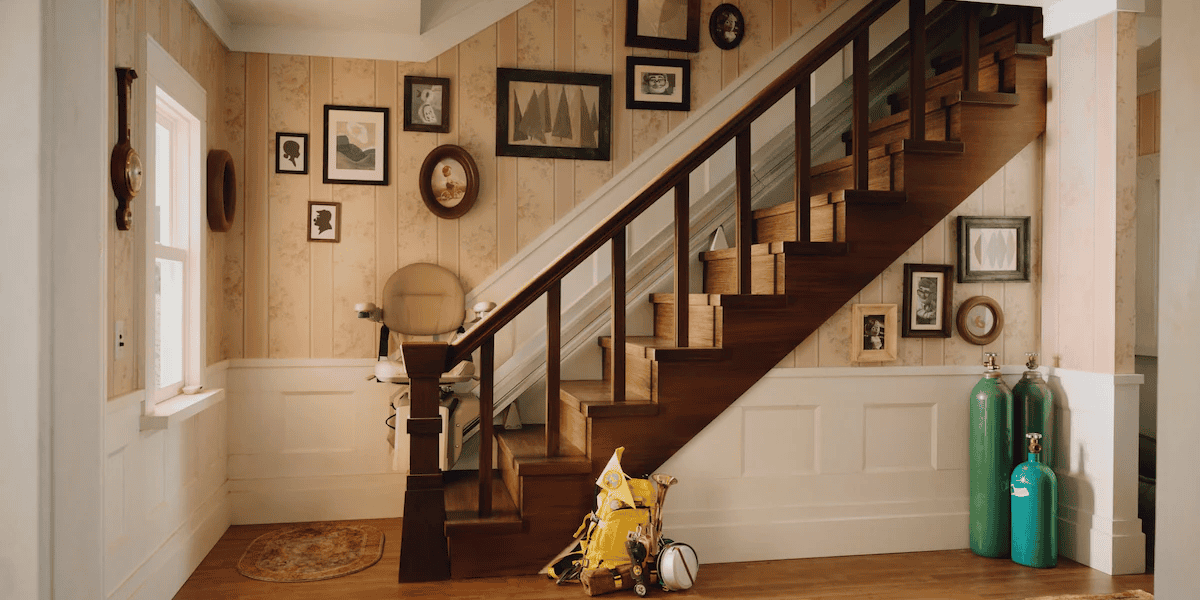 A cozy home interior with a wooden staircase lined with framed photographs and paintings. A vacuum cleaner and two green oxygen tanks rest near the wall.