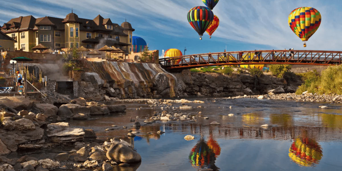 Colorful hot air balloons float above a river with a rocky shoreline, near a bridge connecting to buildings resembling a European-style village, under a clear blue sky.